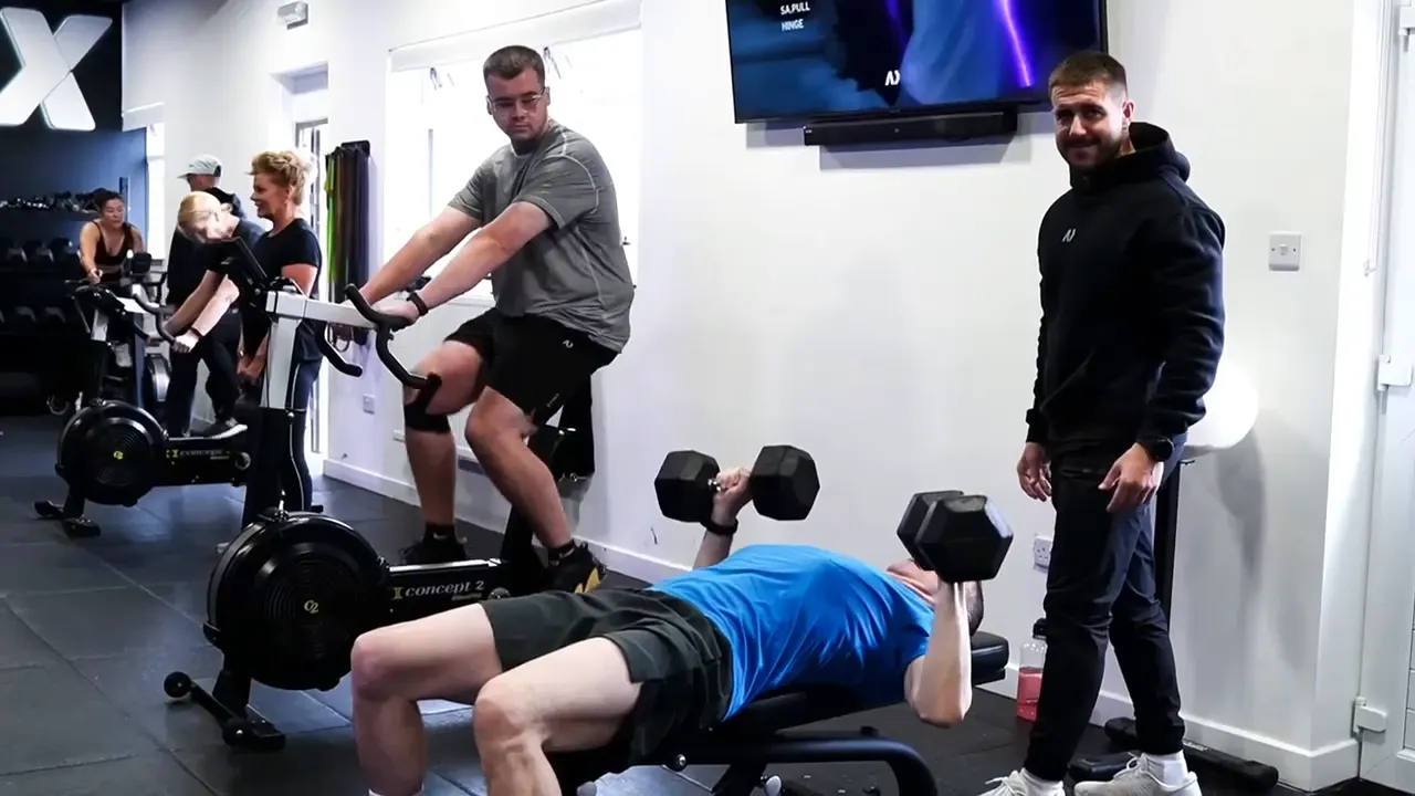 A CMAX coach watching a member's dumbbell press while others train behind